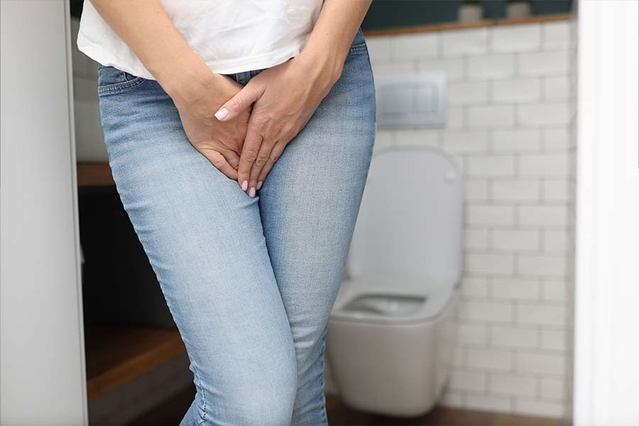 Mulher com as mãos na região da vagina por ter incontinência urinária. Ao fundo, um vaso sanitário e uma parede de tijolos brancos.