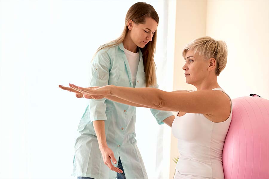 Mulher que teve câncer de mama fazendo fisioterapia para ajudar na recuperação. Ela tem a ajuda de uma fisioterapeuta. O exercício realizado é com um bola e os braços estendidos na altura do ombro. Representação da fisioterapia para câncer de mama.