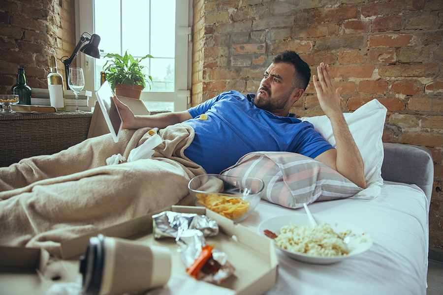 Homem deitado no sofá com várias comidas e bebidas jogadas na cama. O homem está debaixo das cobertas e interagindo com um tablet. Representação da fome emocional.