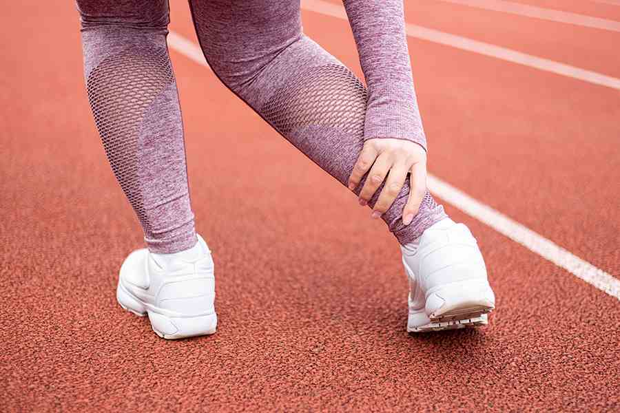 Na pista de corrida, close de pernas de mulher com a mão no tornozelo indicando dor. Representação de lesões mais comuns no esporte.