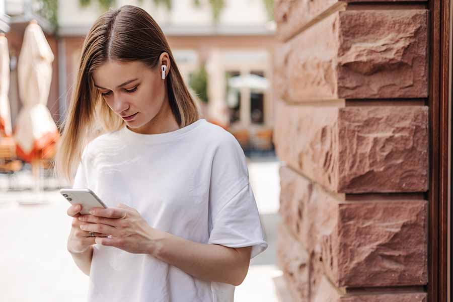 Ao ar livre, mulher mexe no celular e usa fone de ouvidos. Representação dos perigos das rede sociais para a saúde mental.