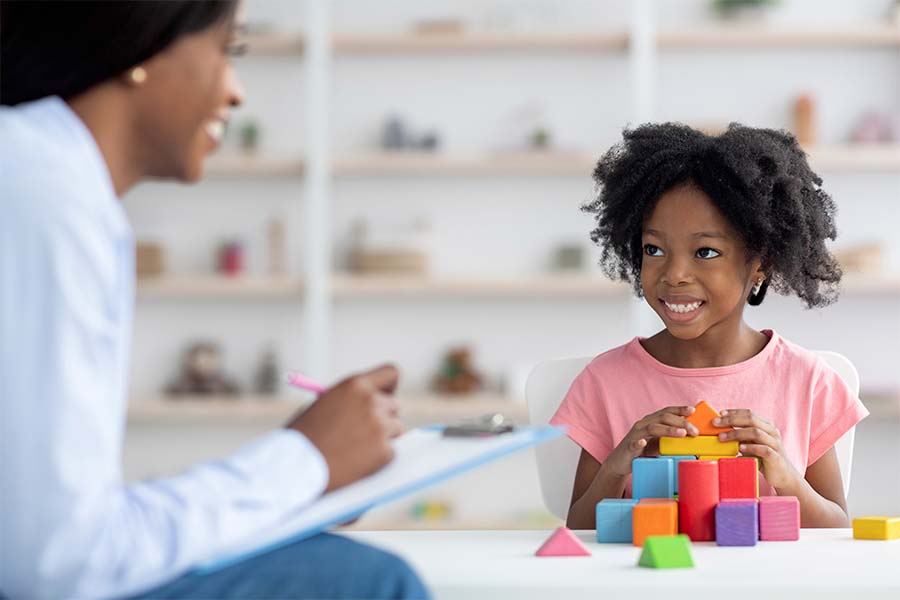 Menina negra em uma sessão de terapia ocupacional com o terapeuta ocupacional também negra. A menina está brincando com blocos coloridos de tamanhos diferentes. Representação de terapia ocupacional para crianças.