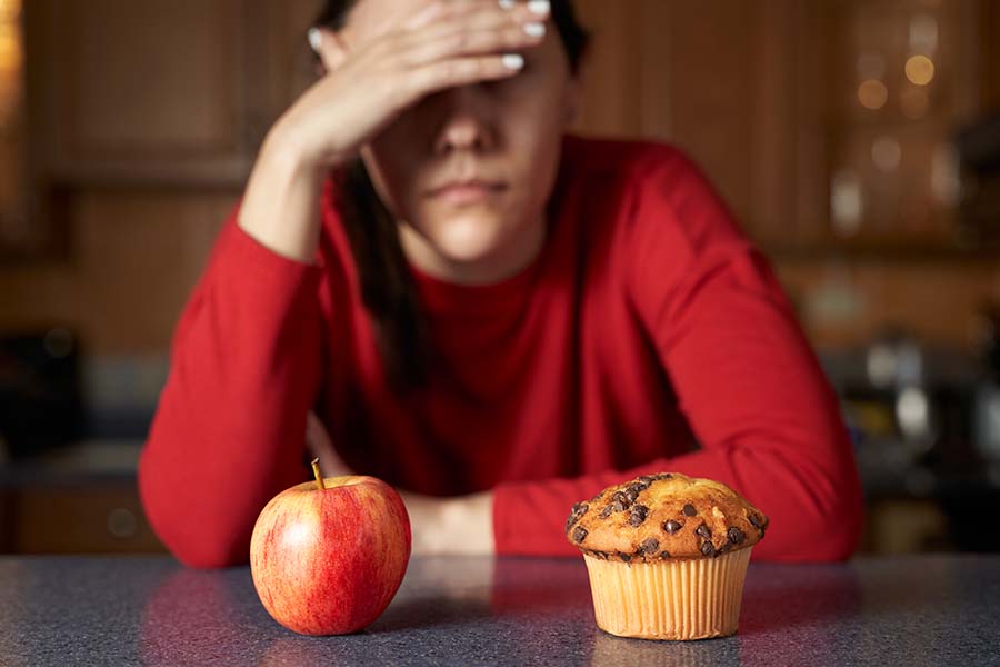 Mulher de vermelho sentada à mesa com uma das mão tampando o rosto. Na frente dela, uma maça e um muffin. Representação dos distúrbios alimentares.