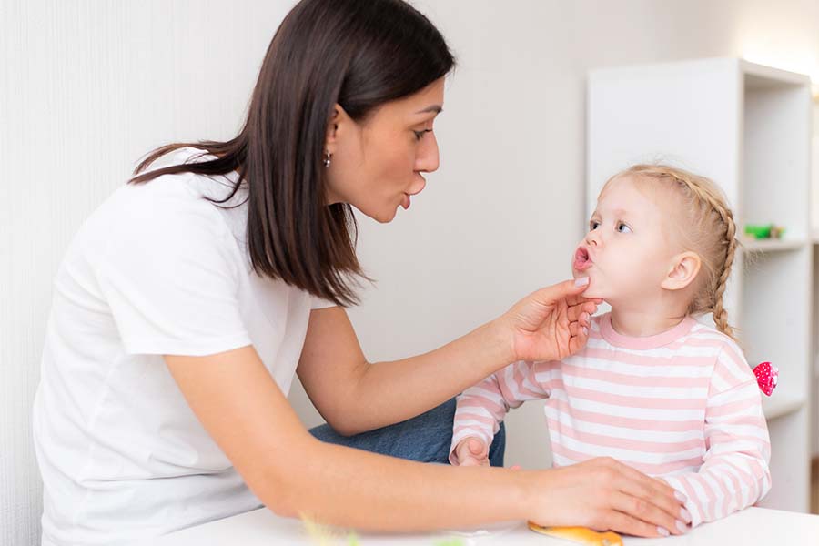 Fonoaudióloga com a mão no queixo de uma paciente criança. A menina está fazendo um biquinho com a boca. Representação dos mitos sobre a fonoaudiologia.