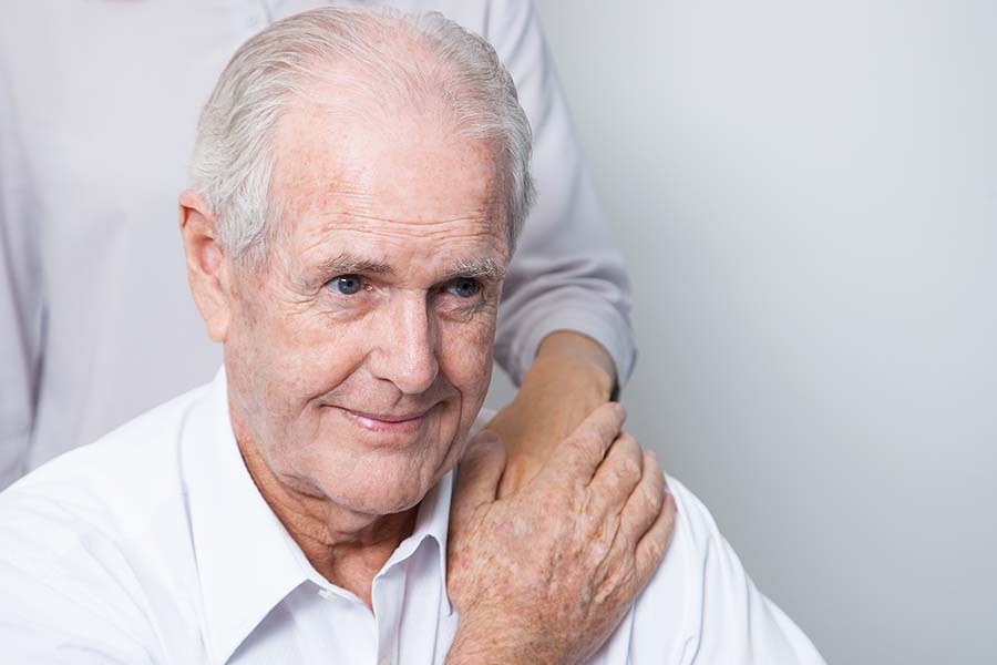 Homem idoso que sofre do mal de Alzheimer segurando a mão de uma mulher que está em pé atrás dele apoiando no seu ombro.