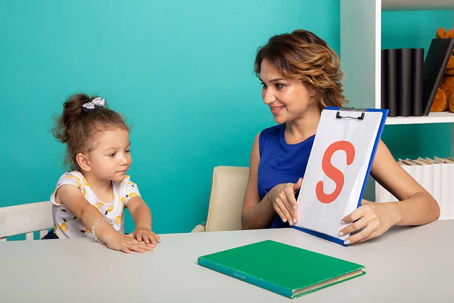 Fonoaudióloga segurando uma placa com a letra S enquanto uma menina pequena reproduz a letra com a voz. Representação da fonoaudiologia infantil.