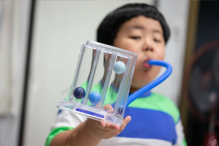 Menino soprando um incentivador respiratório, aparelho com um tubo e 3 bolinhas que se levantam com o sopro do paciente. Representação da fisioterapia para asma.