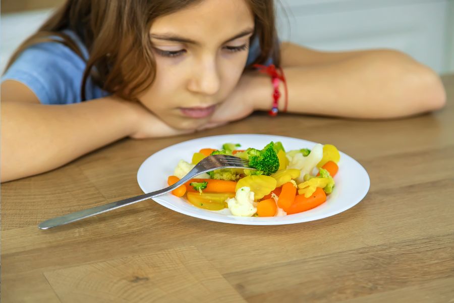 Menina com a cabeça e os braços apoiados na mesa em frente a um prato de vegetais e legumes. Ela está com um olhar triste como se não quisesse comer a comida. Representação de seletividade alimentar.