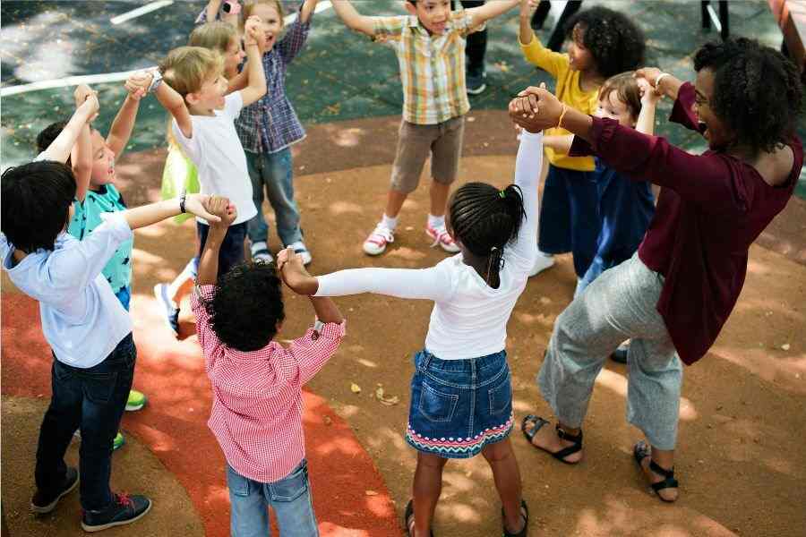 Crianças de mãos dadas em um parque de bairro. Eles estão socializando sorridentes. Tem um adulto no meio deles. Representação de como desenvolver habilidade sociais em crianças.