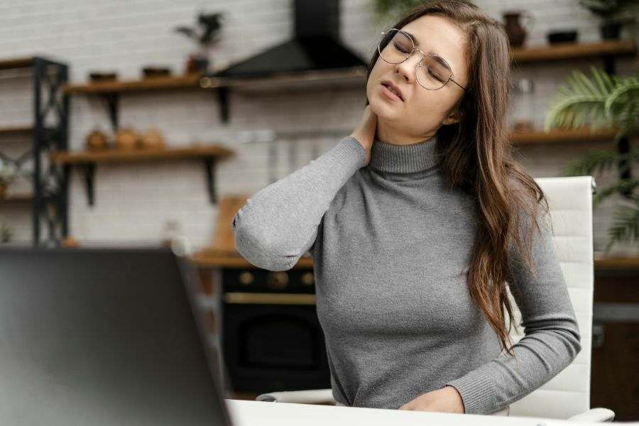 Mulher sentada na mesa da sala de casa na frente de um computador com uma das mãos no pescoço, a cabeça inclinada e o rosto com um semblante que indica dor na região. Representação da dor no pescoço.
