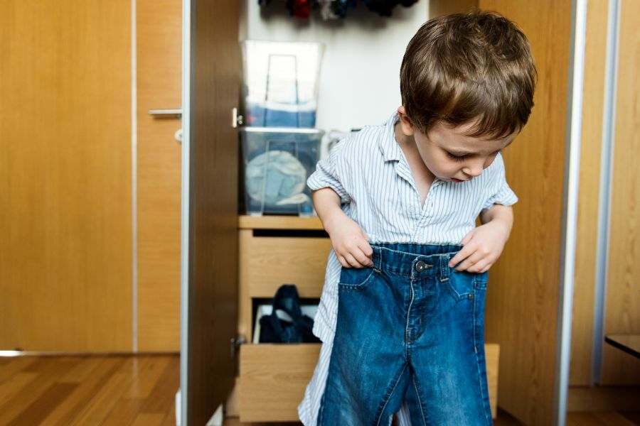 Criança vestindo a própria roupa. Ele está no quarto e atrás tem o armário aberto. Representação da autonomia infantil.