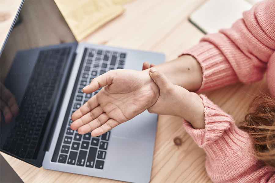 Close de mulher segurando o pulso indicando dor na região. O fundo da imagem é um computador. Representação da lesão por esforço repetitivo, LER.